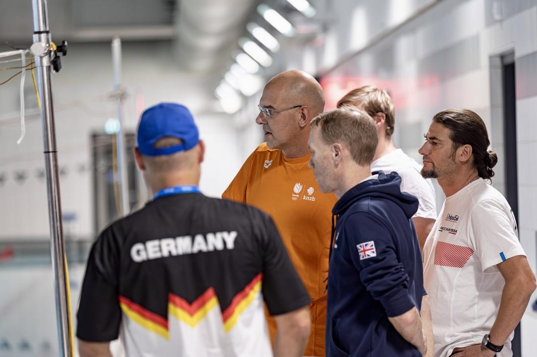 F&uuml;nf Trainer aus verschiedenen Nationen beobachten ihre Athleten im Wasser.