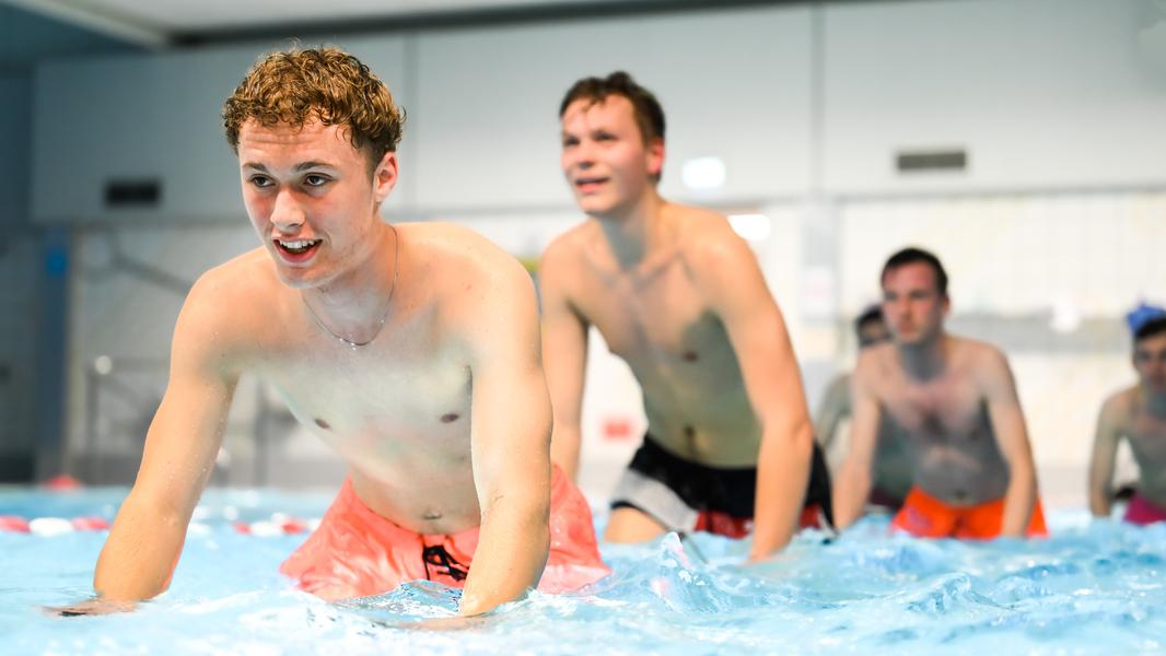 Vier junge Männer in Badebekleidung üben im Wasser eines Schwimmbeckens, einige lachen und spritzen.