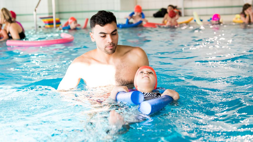 Mann unterstützt ein Kind mit Schwimmhilfen im Wasser eines Schwimmbads, während andere Schwimmer im Hintergrund sind.
